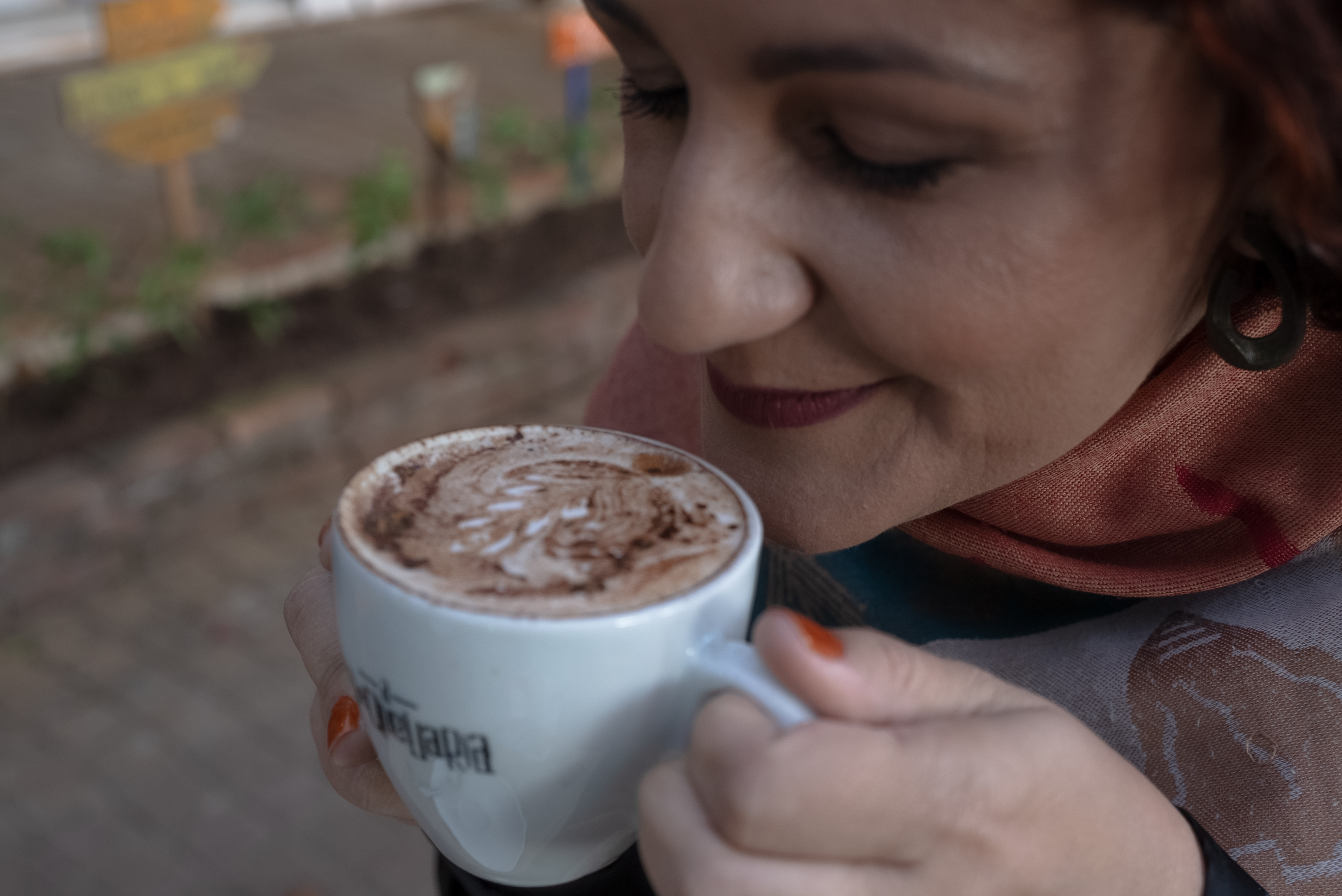 Carol Peixoto com um xale de tons terracota e petróleo, olhando com serenidade para uma xícara de café