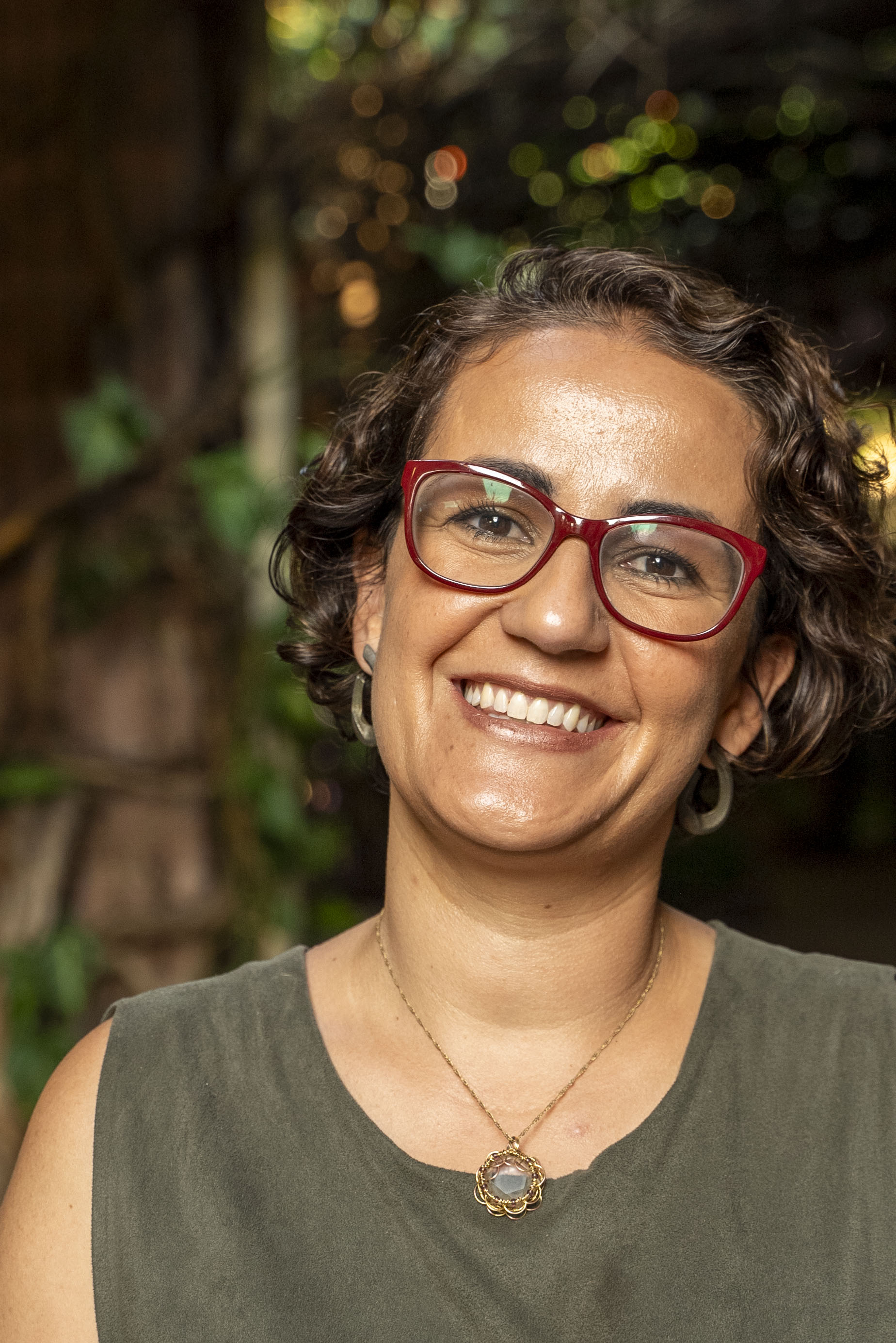 Carol Peixoto sorrindo, com uma xícara de café nas mãos, em um café ao ar livre cercado de plantas