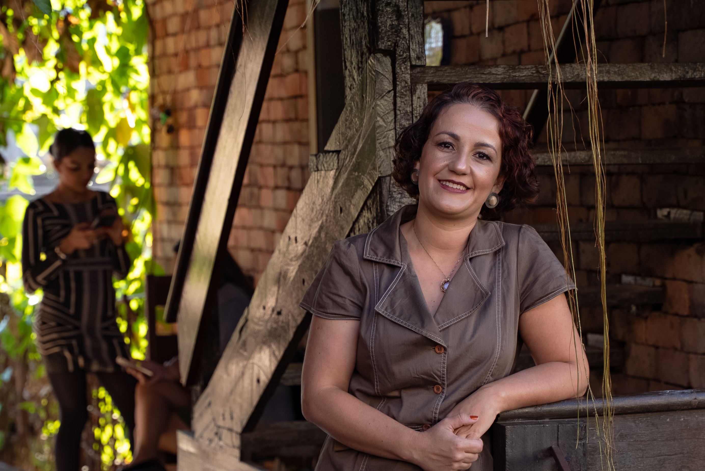 Carol Peixoto sorrindo, encostada em uma escada de madeira rústica, com parede de tijolos e plantas ao fundo